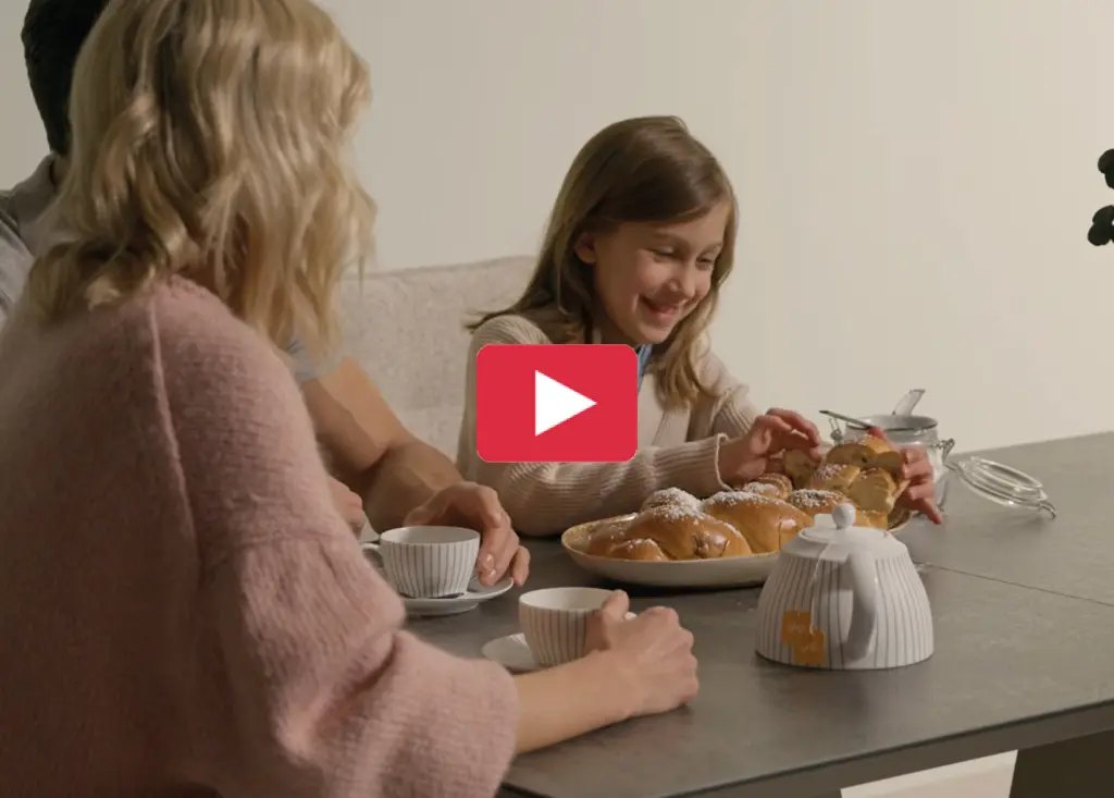 Familie sitzt am Esstisch und genießt Frühstück mit Kaffee und Gebäck. Kind greift nach Brötchen auf einem Teller. Gemütliche Wohnszene im modernen Esszimmer mit Tischplatte in Steinoptik und harmonischer Atmosphäre.